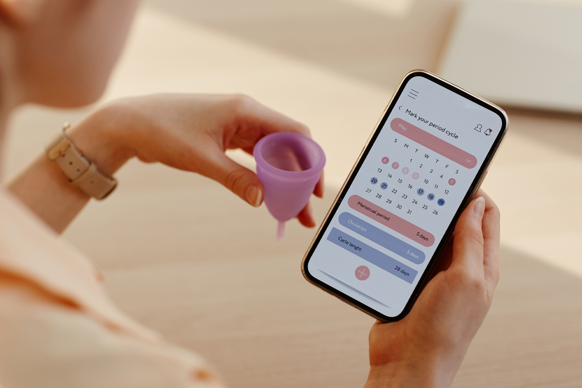 A woman's hands hold a menstrual cup and an iphone showing a fertility monitoring app.