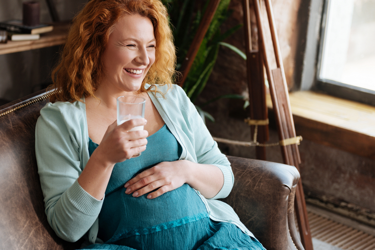 An older pregnant woman smiles while holding a glass of water. 