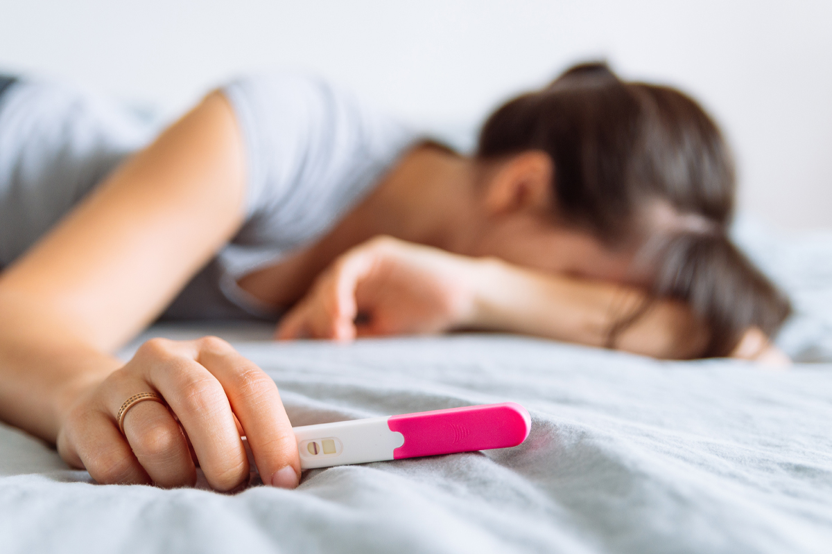 A disappointed woman lays facedown on a bed holding a pregnancy test.