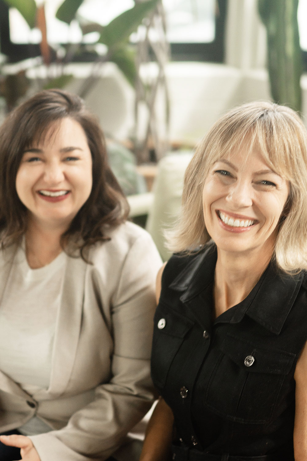 Naturopaths, Fertility Practitioners, and Founders of Positivity Fertility, Dr. Mandy and Dr. Jeanie, smile at each other on a couch. 