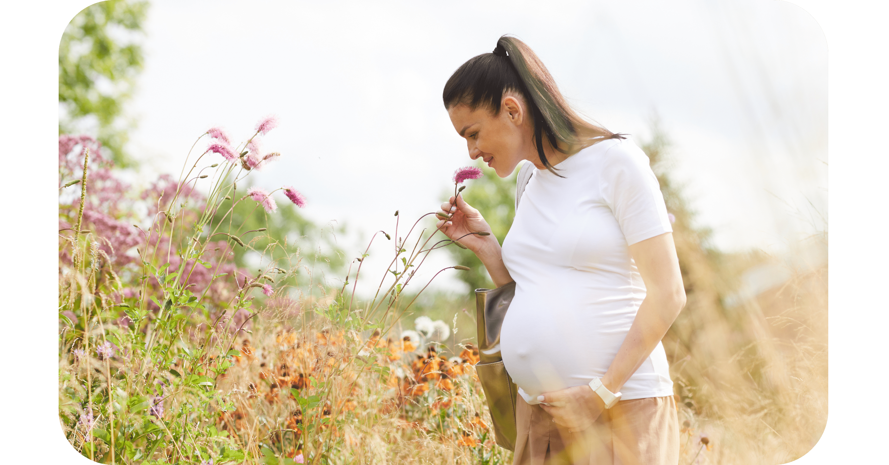 A pregnant woman on a natural fertility journey smiles while smelling a flower in a beautiful field of blossoms.